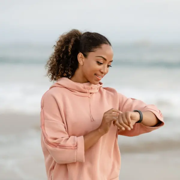 Smiling woman in a pink hoodie checking her smartwatch at the beach